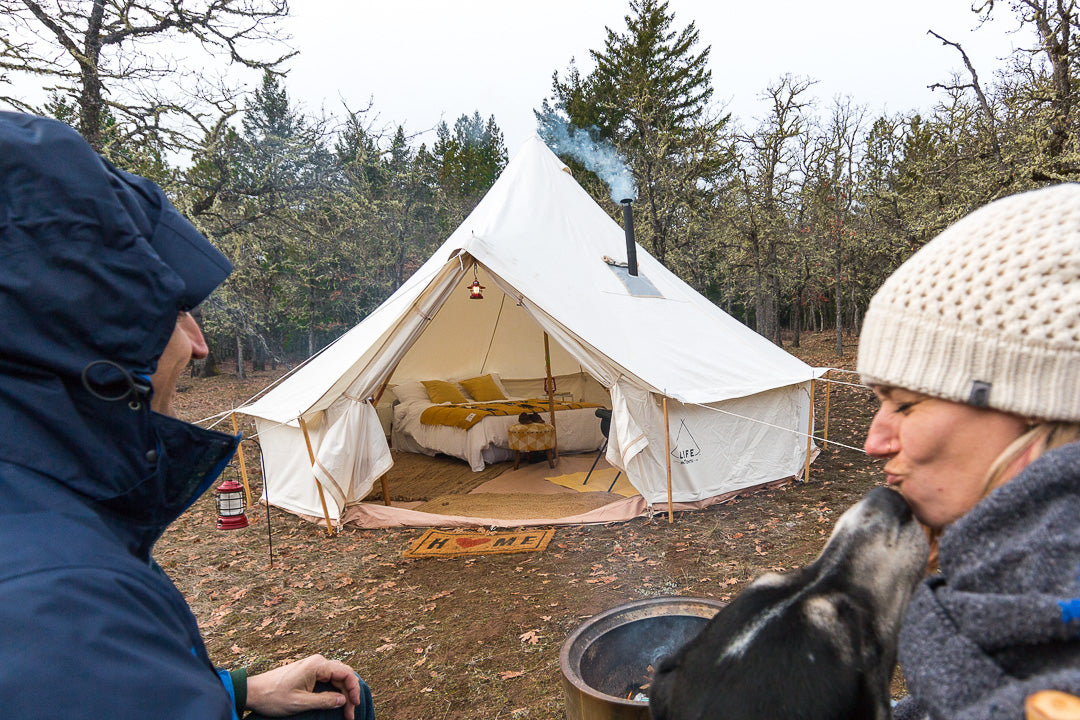 two people and a dog sit outside a canvas tent that has a wood stove piping smoke out the top
